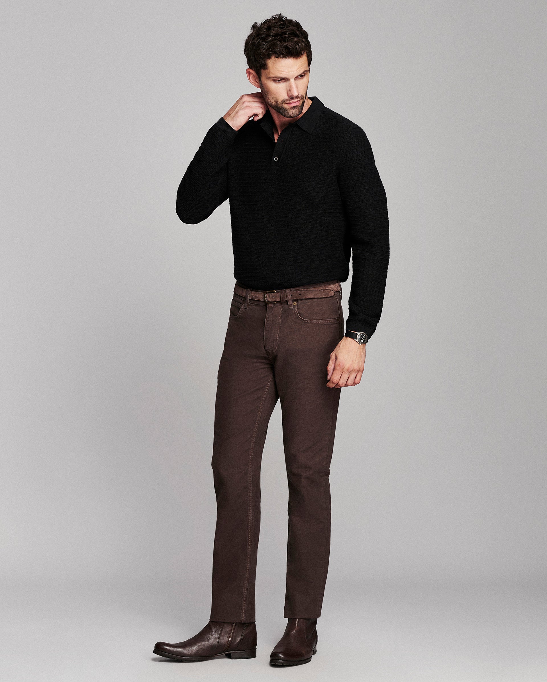 A man modeling a black ribbed knit polo shirt and dark brown trousers with brown leather boots, standing against a solid light gray studio background.