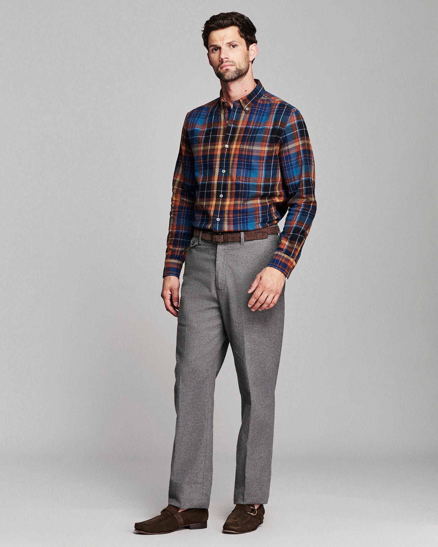 A man modeling a long-sleeve button-down plaid shirt in blue, orange, and red, paired with gray trousers, a brown belt, and brown loafers, against a solid light gray background.