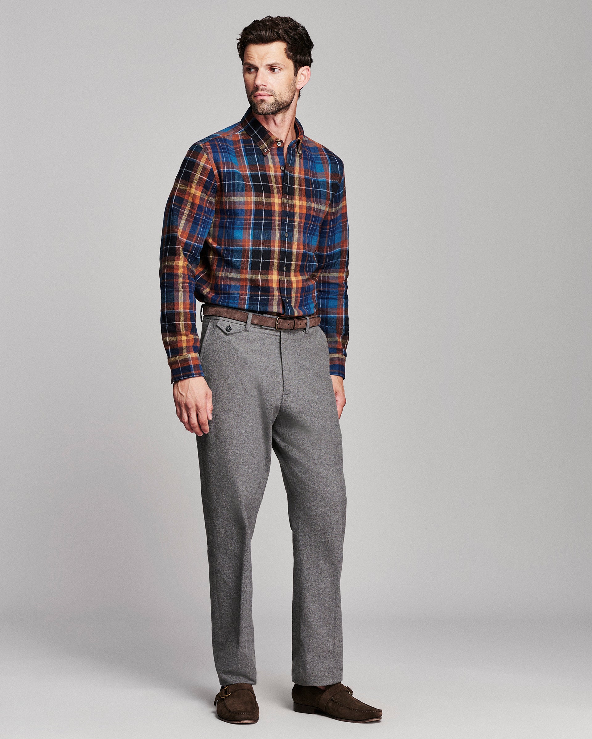 A man modeling a long-sleeve plaid button-down shirt in blue, orange, and brown, paired with gray trousers and brown suede shoes, against a solid gray studio background.