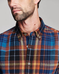Close-up of a man wearing a button-down plaid shirt in blue, orange, black, and beige tones, showing the collar, buttons, and fabric texture against a plain light gray background.