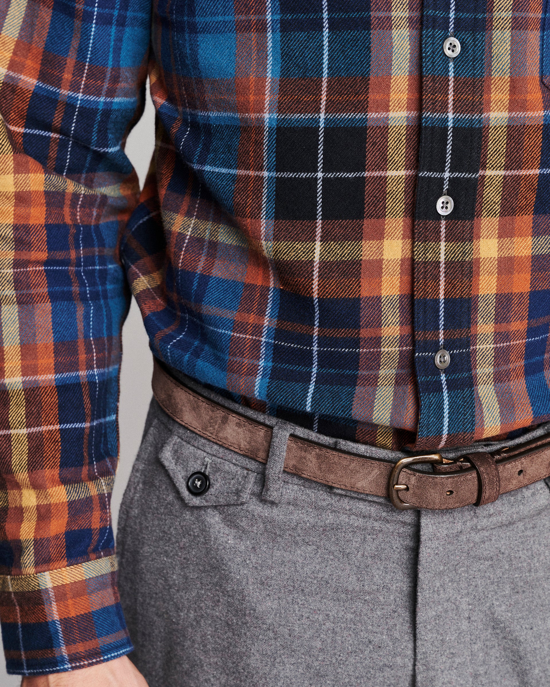 A close-up of a person wearing a long-sleeve plaid button-up shirt in blue, orange, black, and beige tones, tucked into gray trousers with a brown belt.