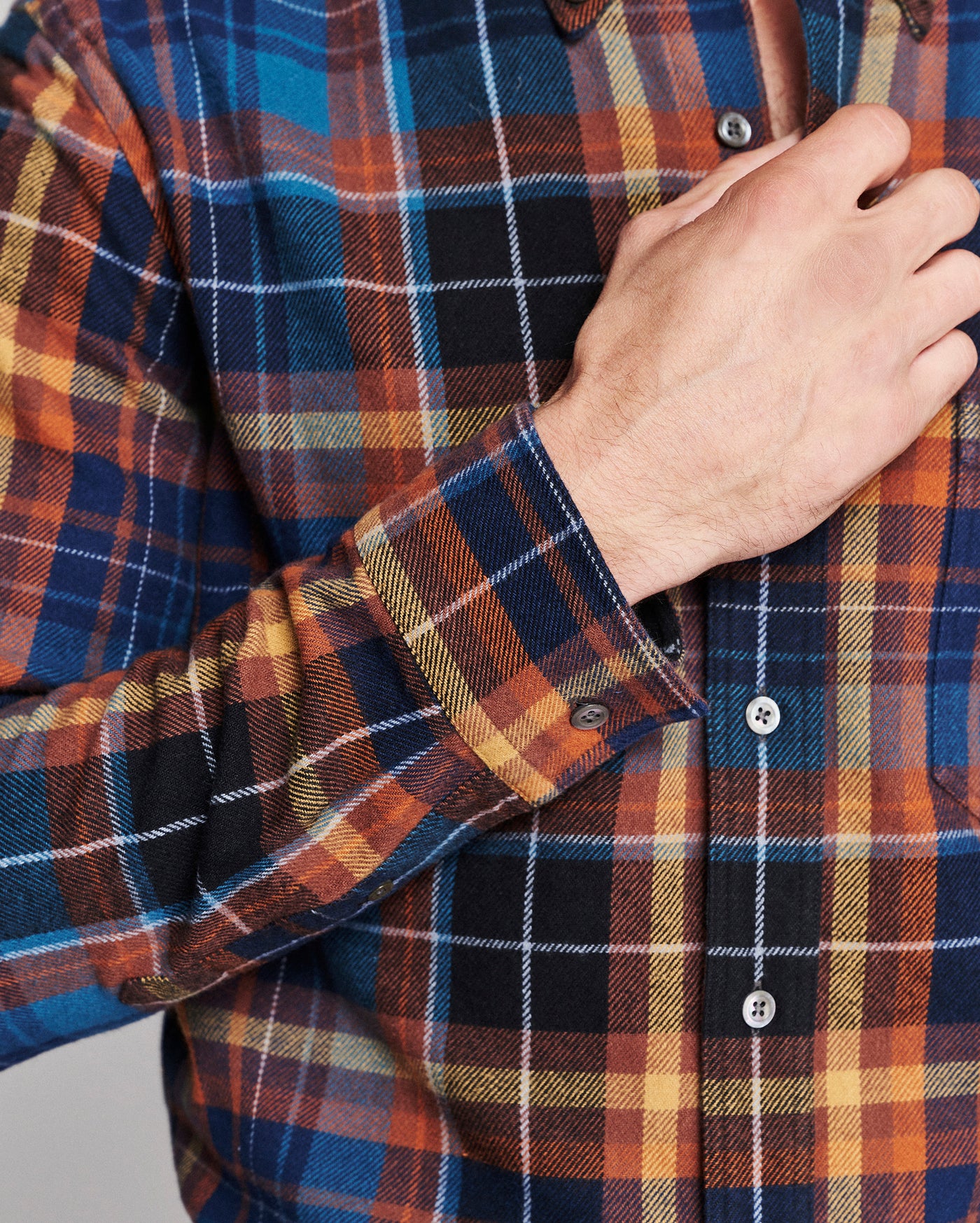 Close-up of a man wearing a long-sleeve plaid button-up shirt, showing his hand adjusting the collar and cuff. The shirt features a multi-colored plaid pattern with blue, orange, black, and yellow tones.