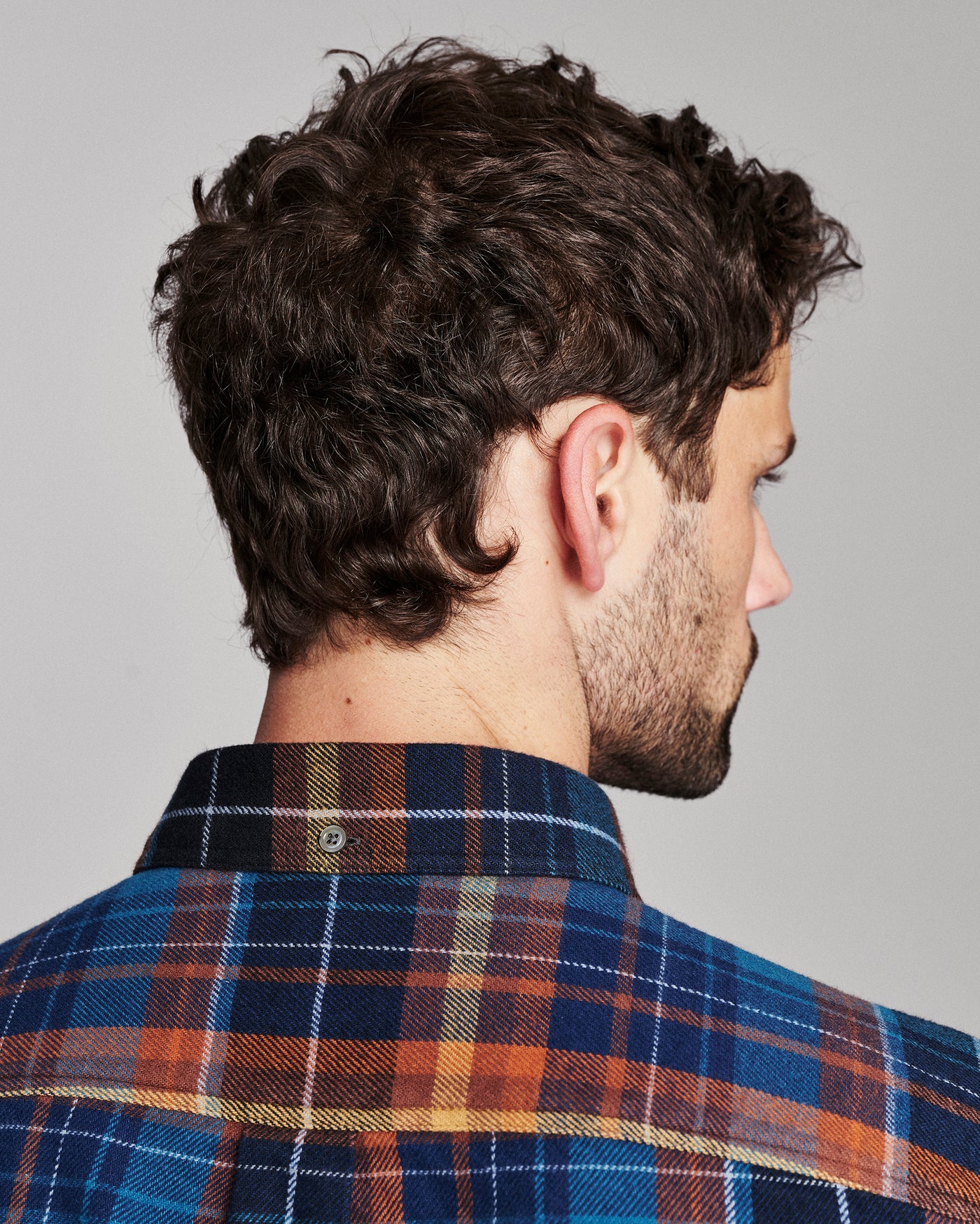 A man shown from the back and side profile wearing a blue, orange, and yellow plaid flannel shirt against a solid gray background.