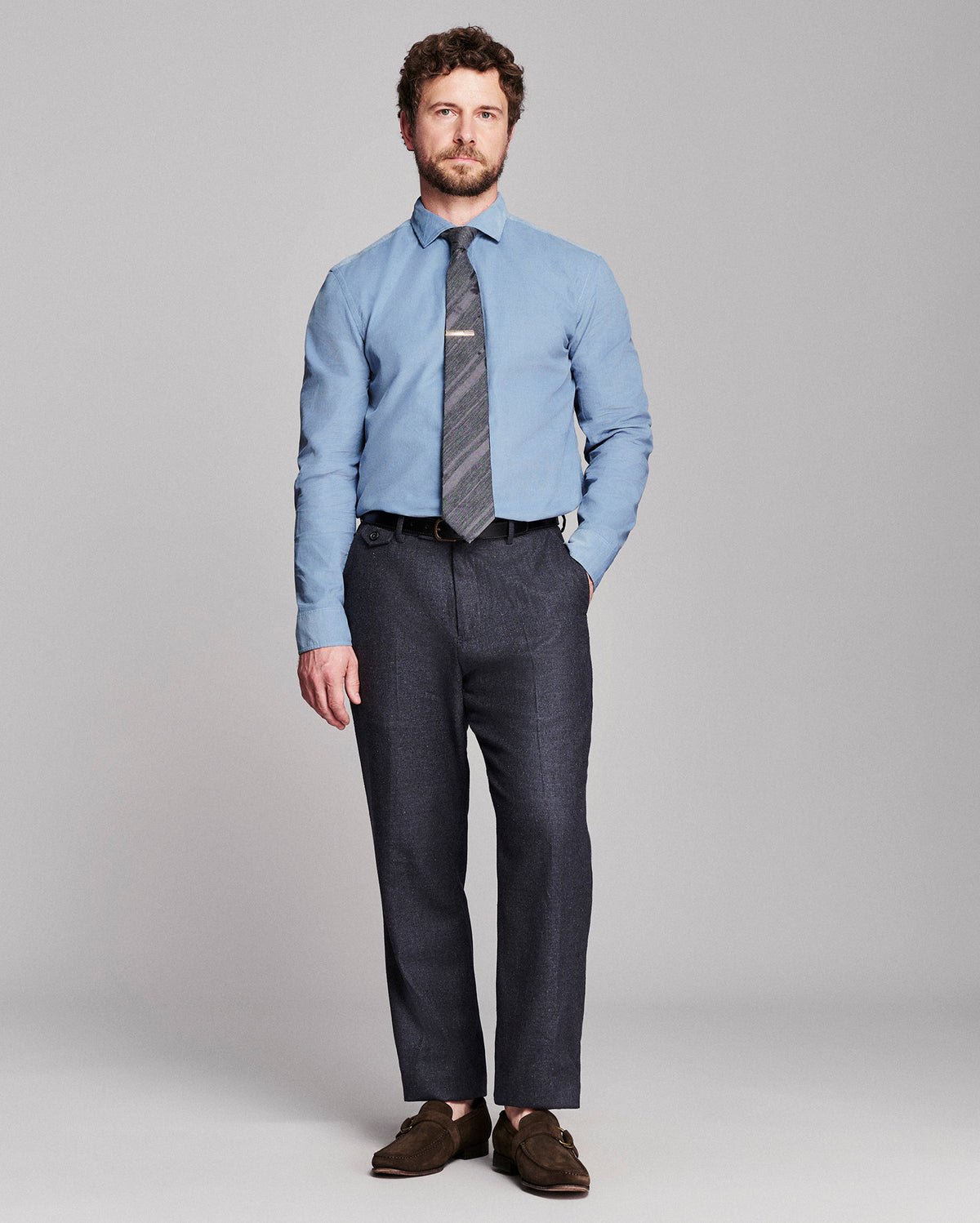 A man modeling a light blue dress shirt, a gray patterned tie, dark charcoal trousers, and brown suede loafers against a solid gray studio background.