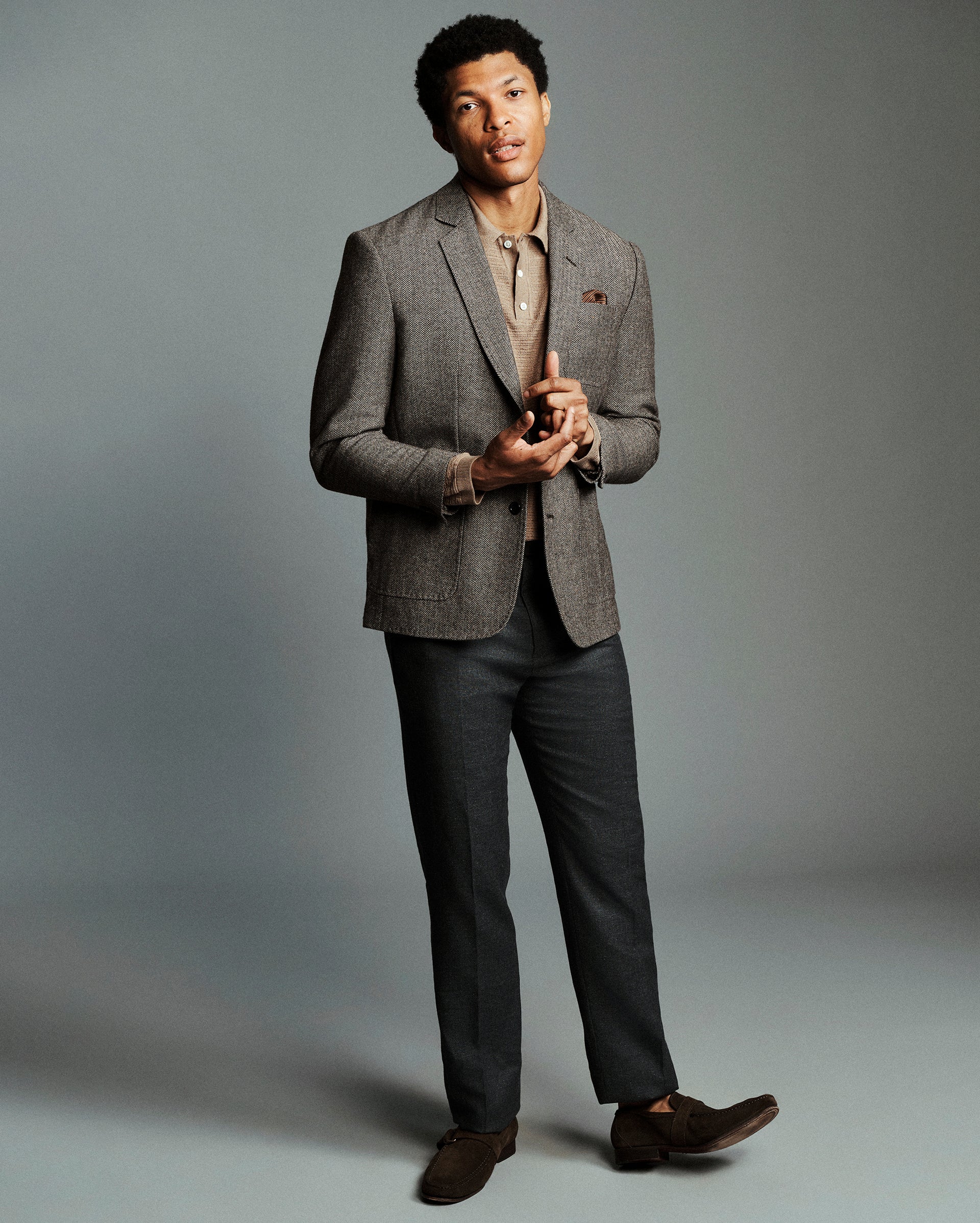 A man modeling a brown herringbone blazer over a beige shirt, paired with dark gray trousers and brown leather shoes, against a solid gray studio background.