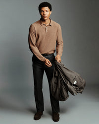A man stands against a gray studio background wearing a tan ribbed knit polo shirt, dark trousers, and brown shoes, while holding a dark gray jacket.
