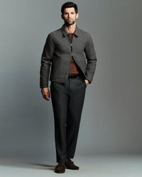 A man modeling a dark gray textured zip-up jacket over a brown shirt, paired with dark gray trousers and brown suede loafers, against a solid gray studio background.