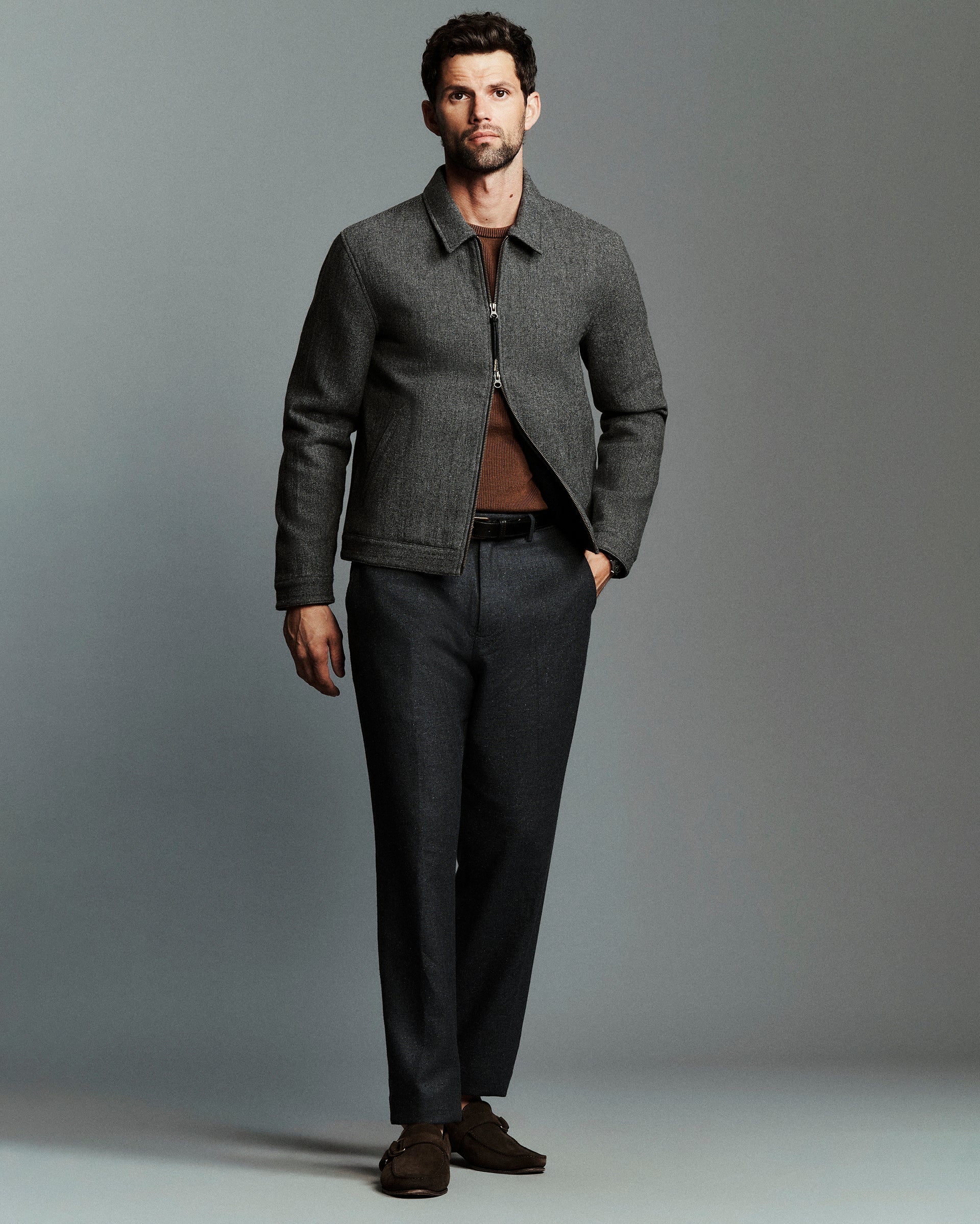 A man modeling a dark gray textured zip-up jacket over a brown shirt, paired with dark gray trousers and brown suede loafers, against a solid gray studio background.