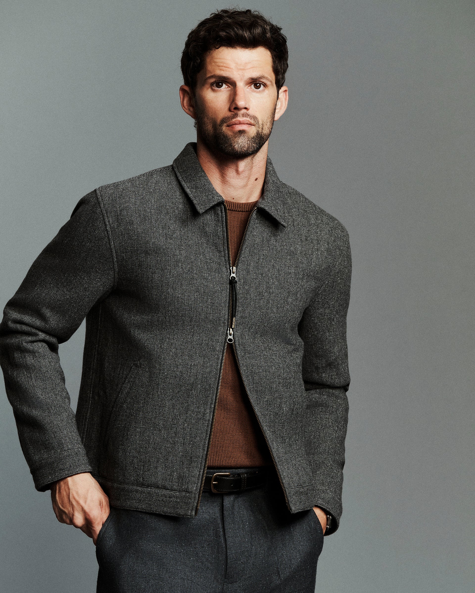 A man wearing a dark gray textured zip-up jacket over a brown shirt and gray trousers, standing against a solid gray studio background.