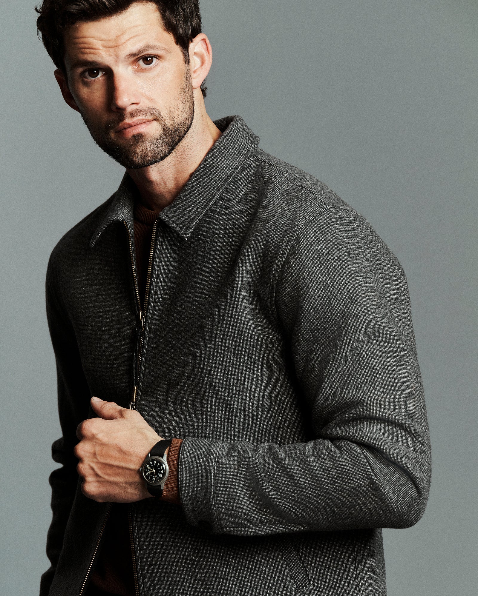 A man wearing a dark gray textured zip-up jacket and a black wristwatch, posed against a solid gray studio background.