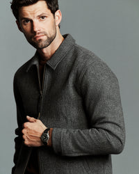 A man wearing a dark gray textured zip-up jacket and a black wristwatch, posed against a solid gray studio background.