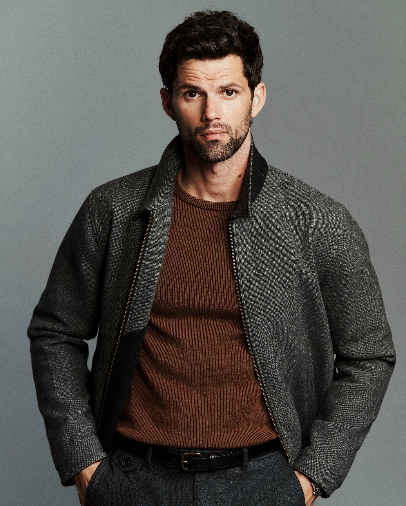 A man wearing a gray textured zip-up jacket with black ribbed trim over a brown knit sweater, photographed against a solid gray studio background.
