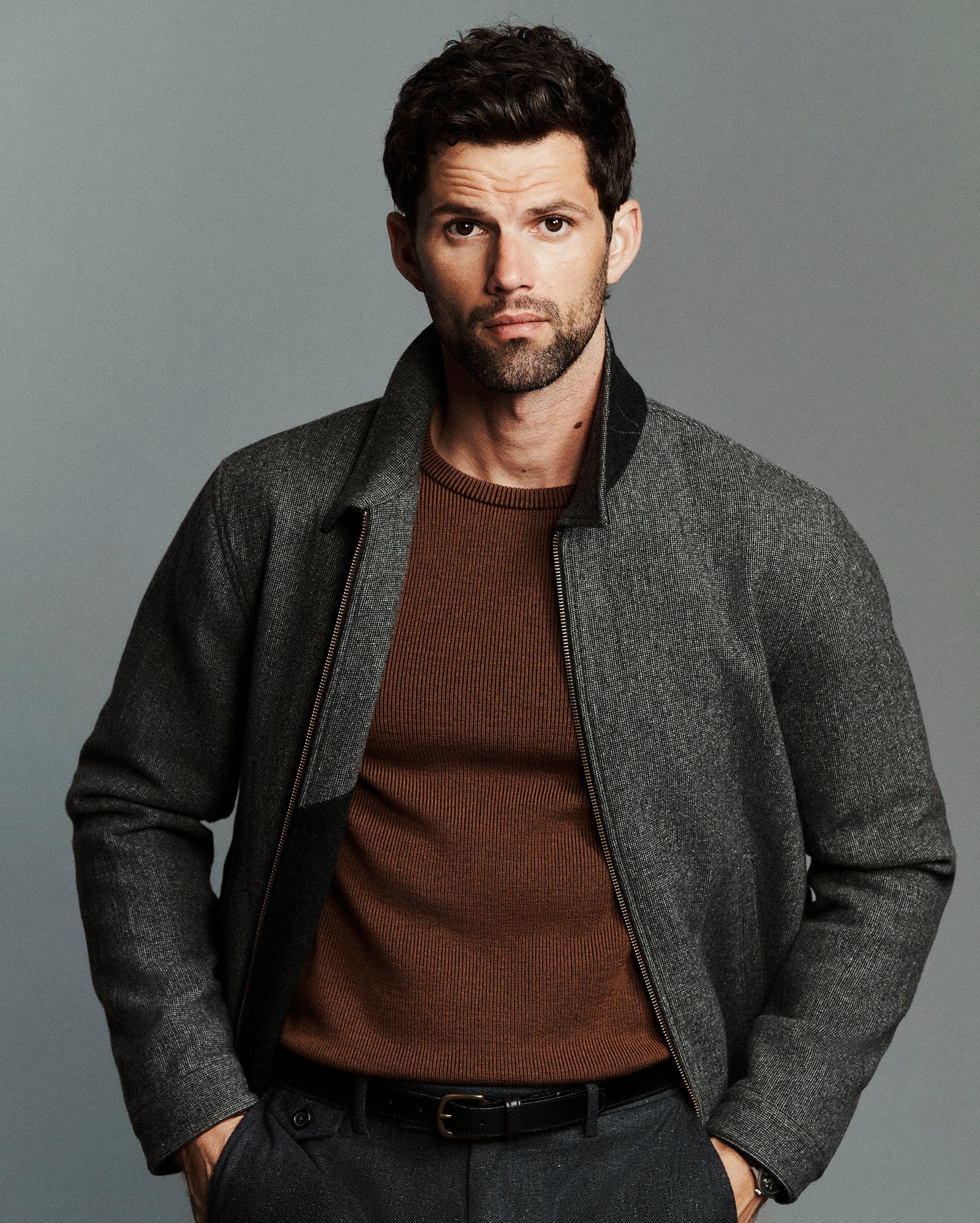 A man wearing a gray textured zip-up jacket with black ribbed trim over a brown knit sweater, photographed against a solid gray studio background.