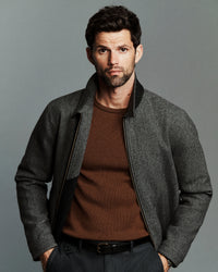 A man wearing a gray textured zip-up jacket with black ribbed trim over a brown knit sweater, photographed against a solid gray studio background.