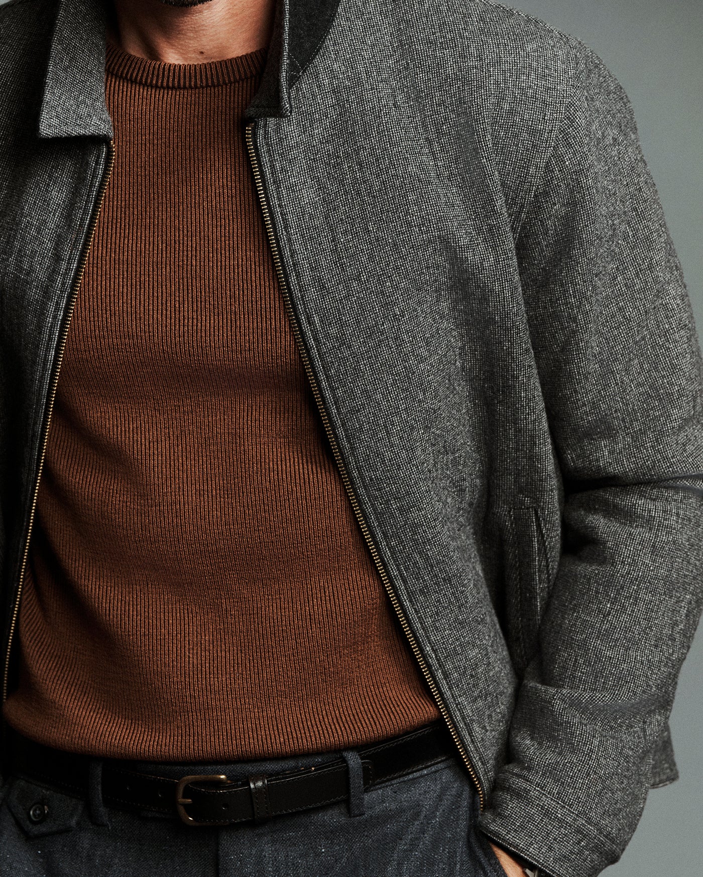 A close-up of a man wearing a dark gray textured zip-up jacket over a brown ribbed knit sweater, paired with a black belt and dark trousers.