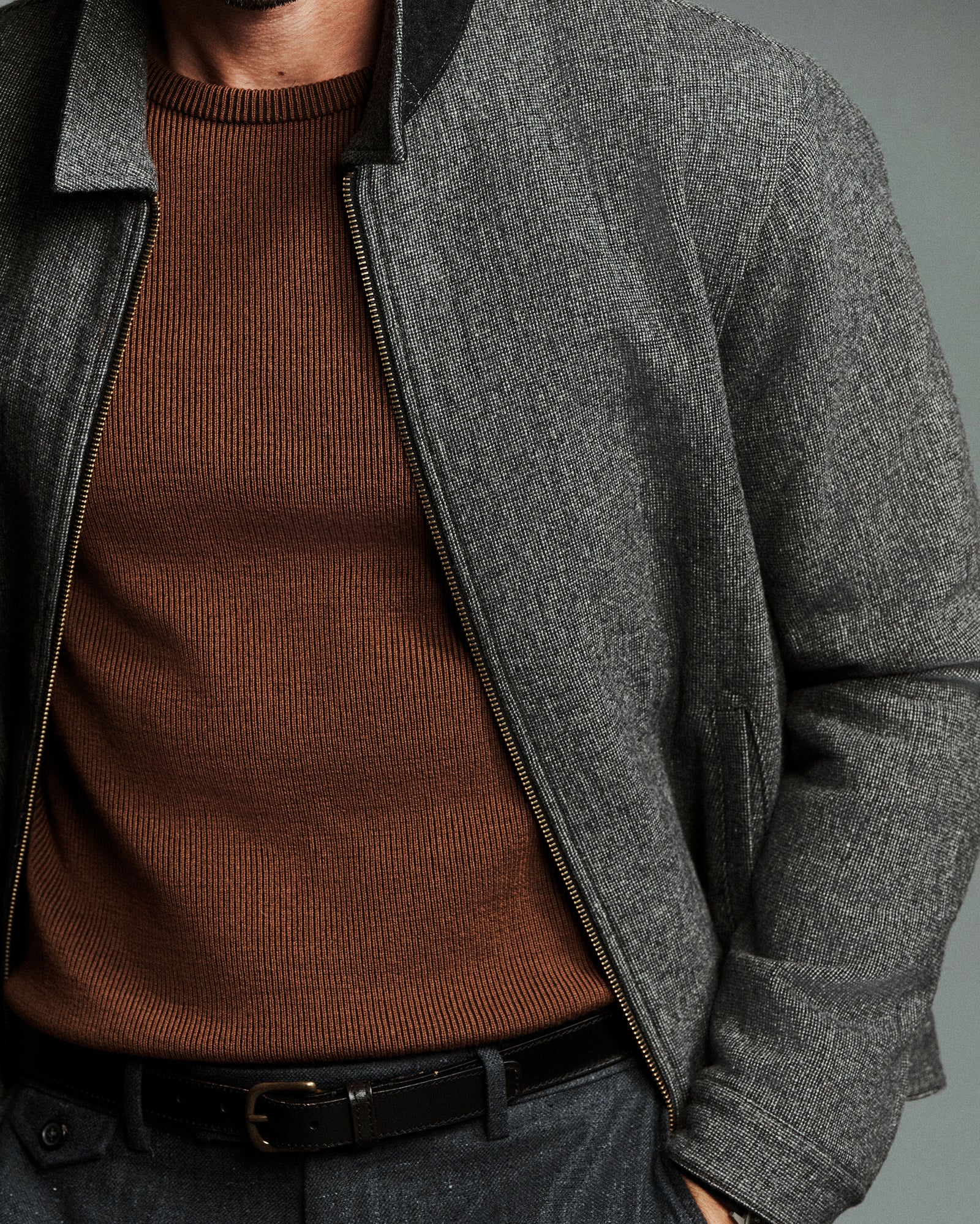 A close-up of a man wearing a dark gray textured zip-up jacket over a brown ribbed knit sweater, paired with a black belt and dark trousers.