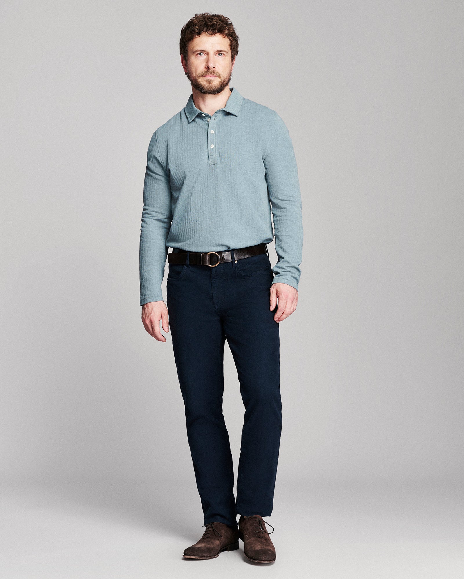 A man modeling a light blue long-sleeve knit polo shirt with dark navy pants and brown leather shoes, standing against a neutral gray studio background.
