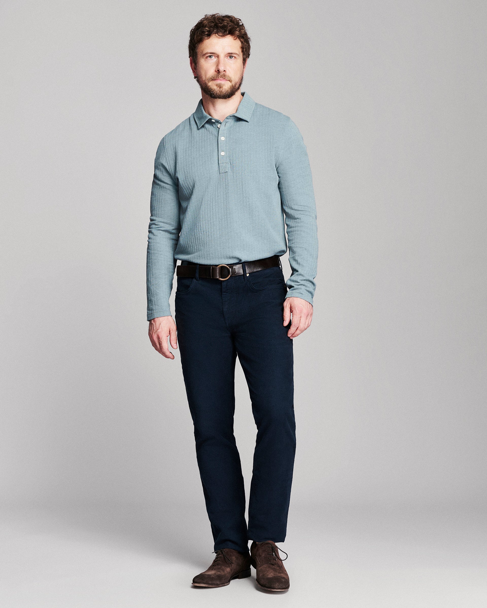 A man modeling a light blue long-sleeve knit polo shirt with dark navy pants and brown leather shoes, standing against a neutral gray studio background.