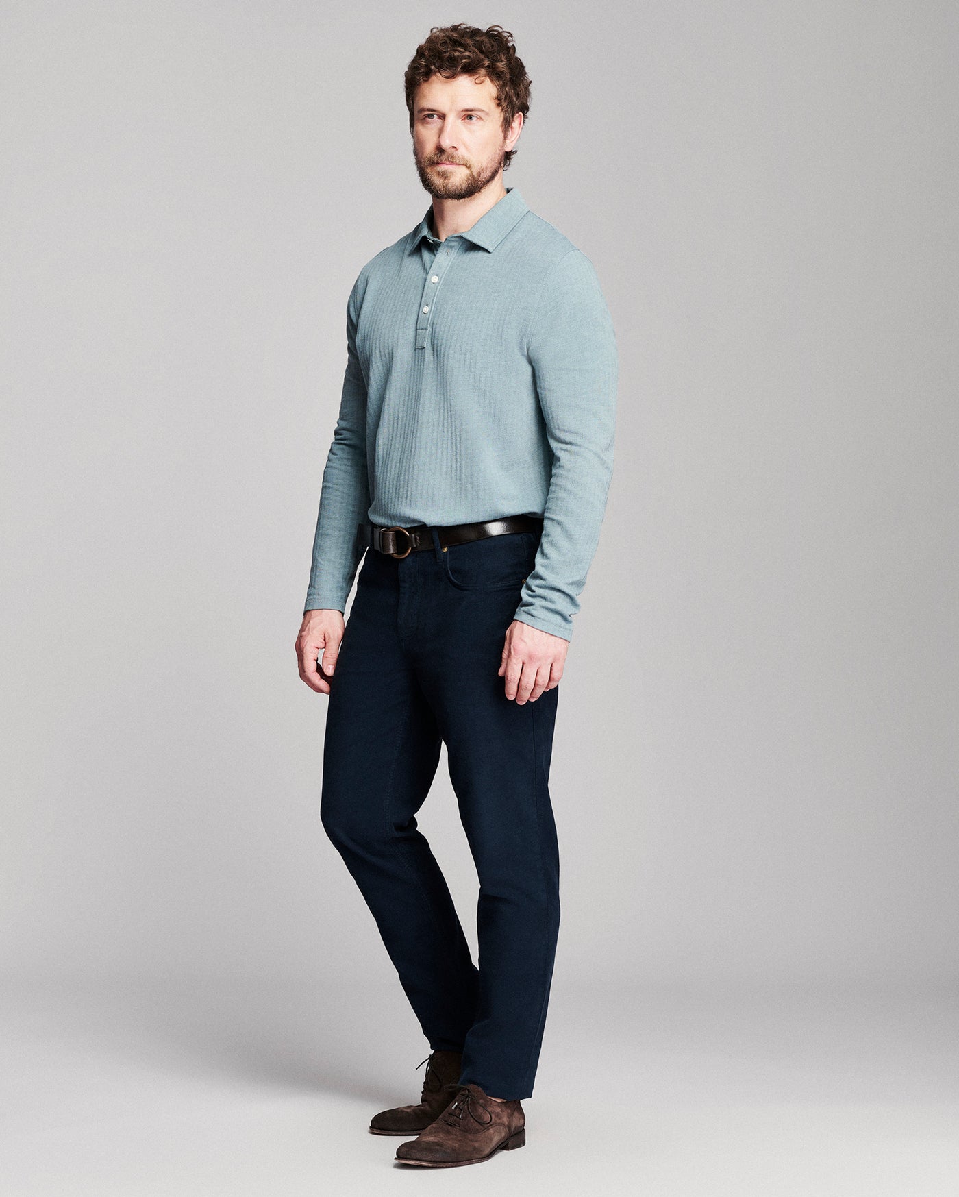 A man modeling a light blue ribbed long-sleeve polo shirt with dark navy trousers, a black belt, and brown suede shoes, standing against a neutral gray studio background.