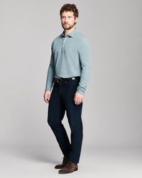 A man modeling a light blue ribbed long-sleeve polo shirt with dark navy trousers, a black belt, and brown suede shoes, standing against a neutral gray studio background.