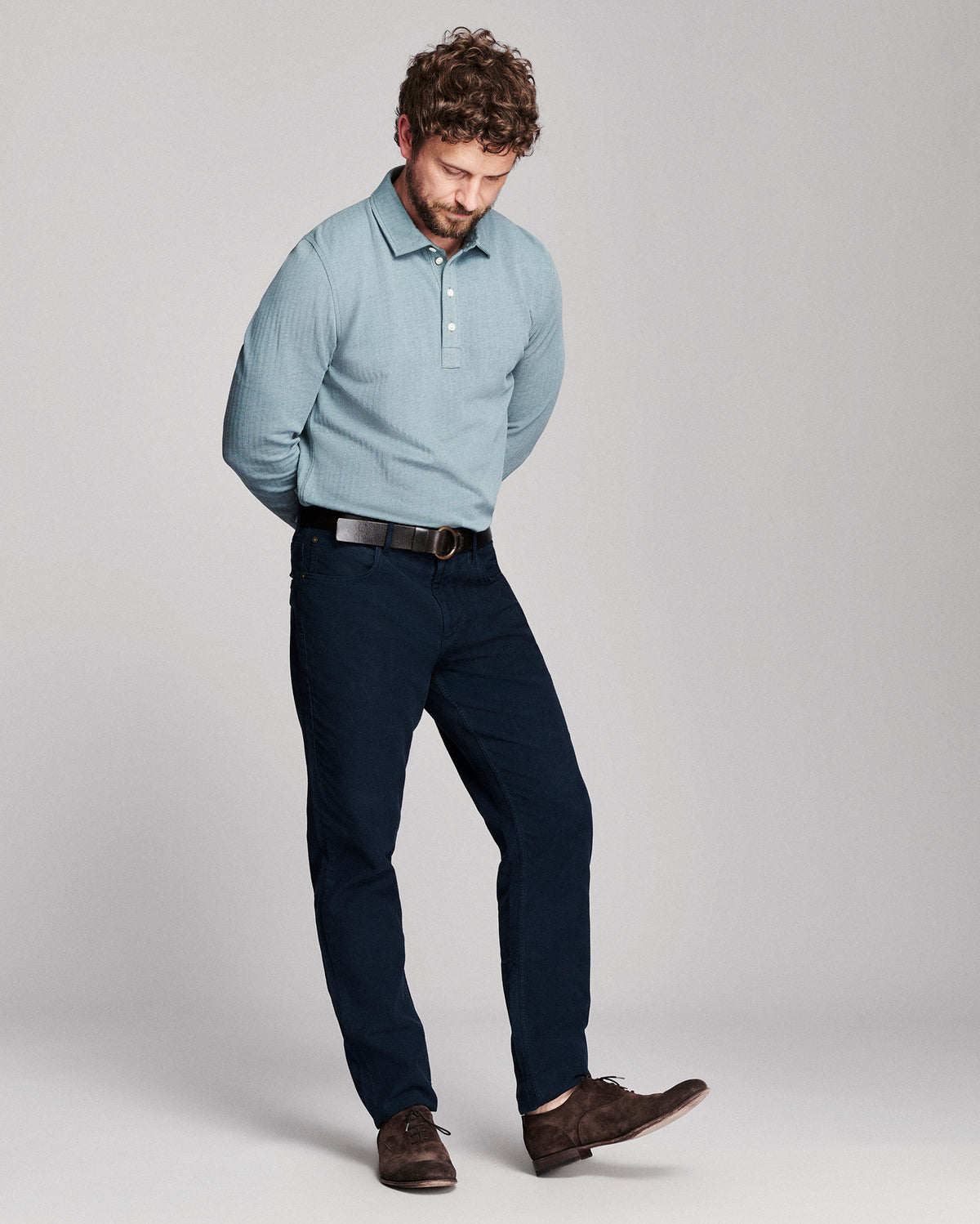 A man modeling a light blue long-sleeve polo shirt and dark navy trousers, paired with a brown belt and brown suede shoes, against a neutral gray studio background.