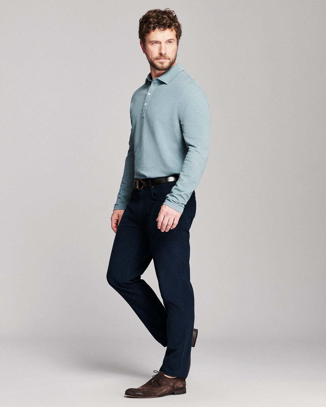 A man modeling a light blue long-sleeve knit polo shirt with dark navy trousers and brown leather shoes, standing against a neutral gray studio background.