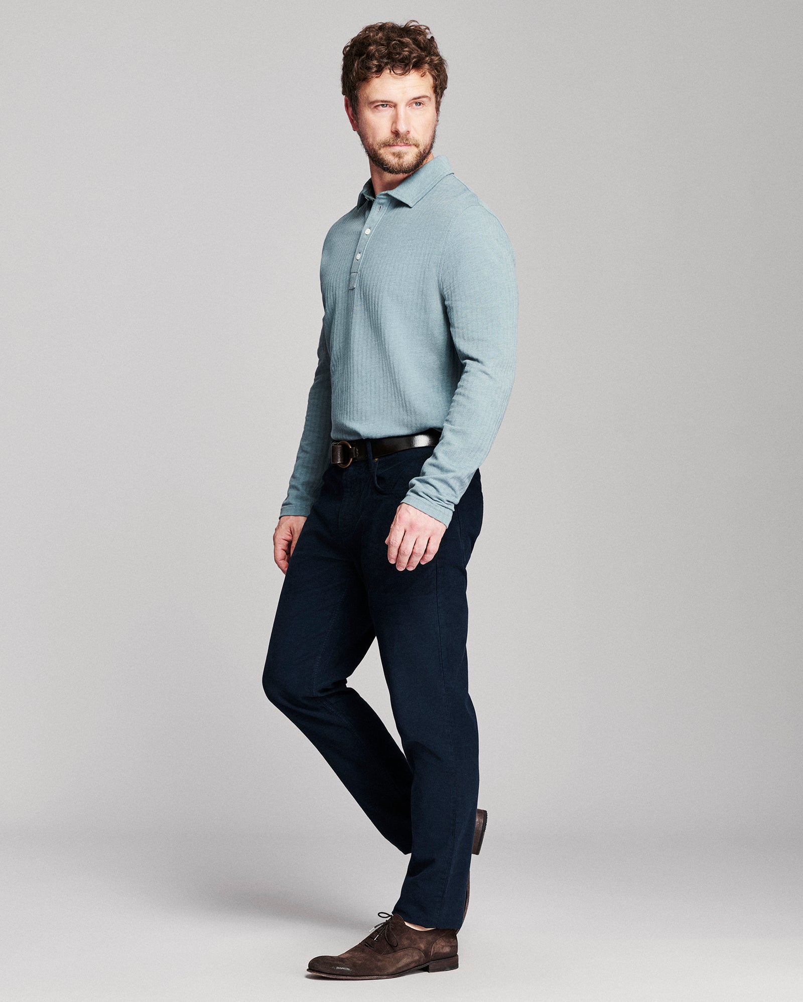 A man modeling a light blue long-sleeve knit polo shirt with dark navy trousers and brown leather shoes, standing against a neutral gray studio background.