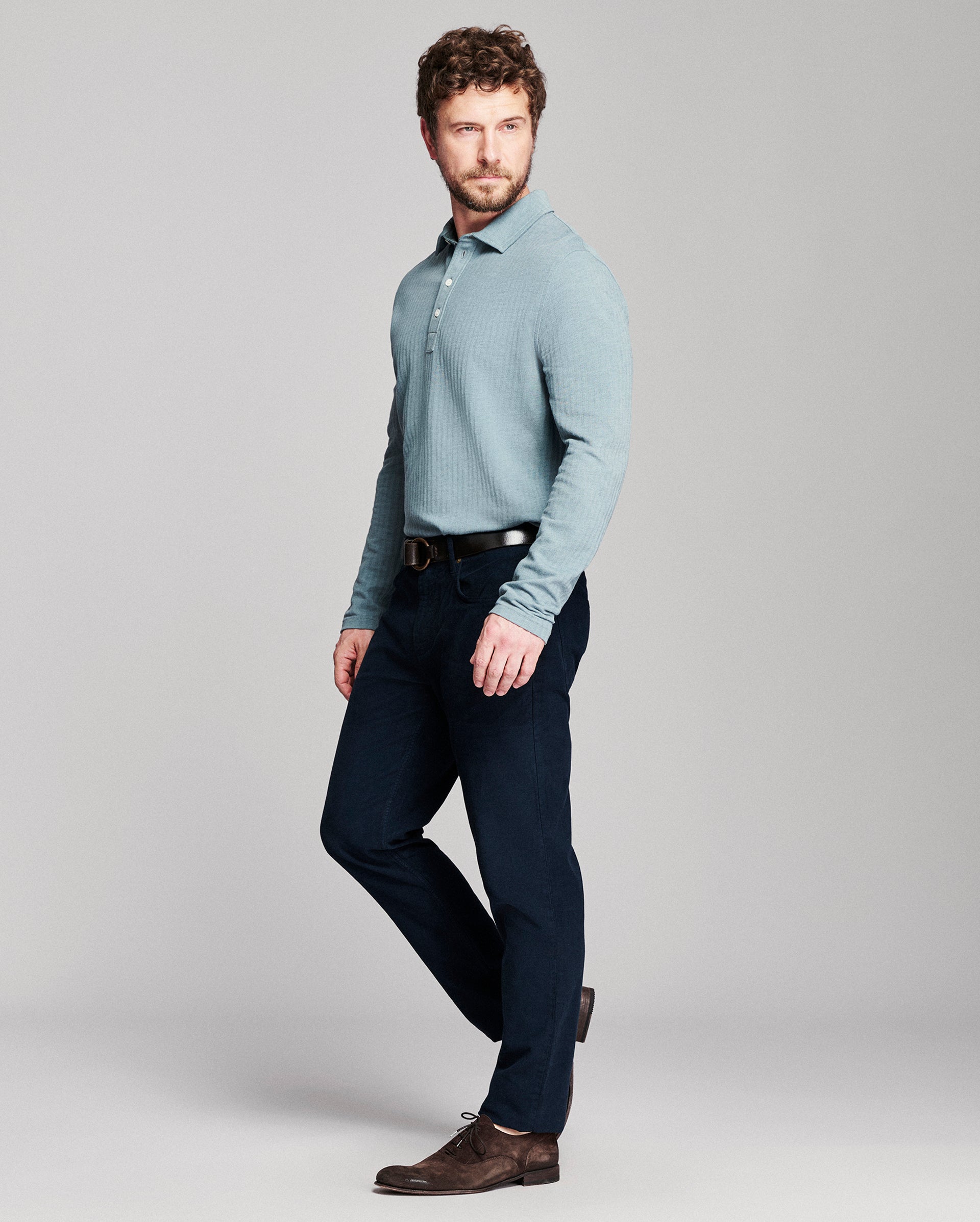 A man modeling a light blue long-sleeve knit polo shirt with dark navy trousers and brown leather shoes, standing against a neutral gray studio background.