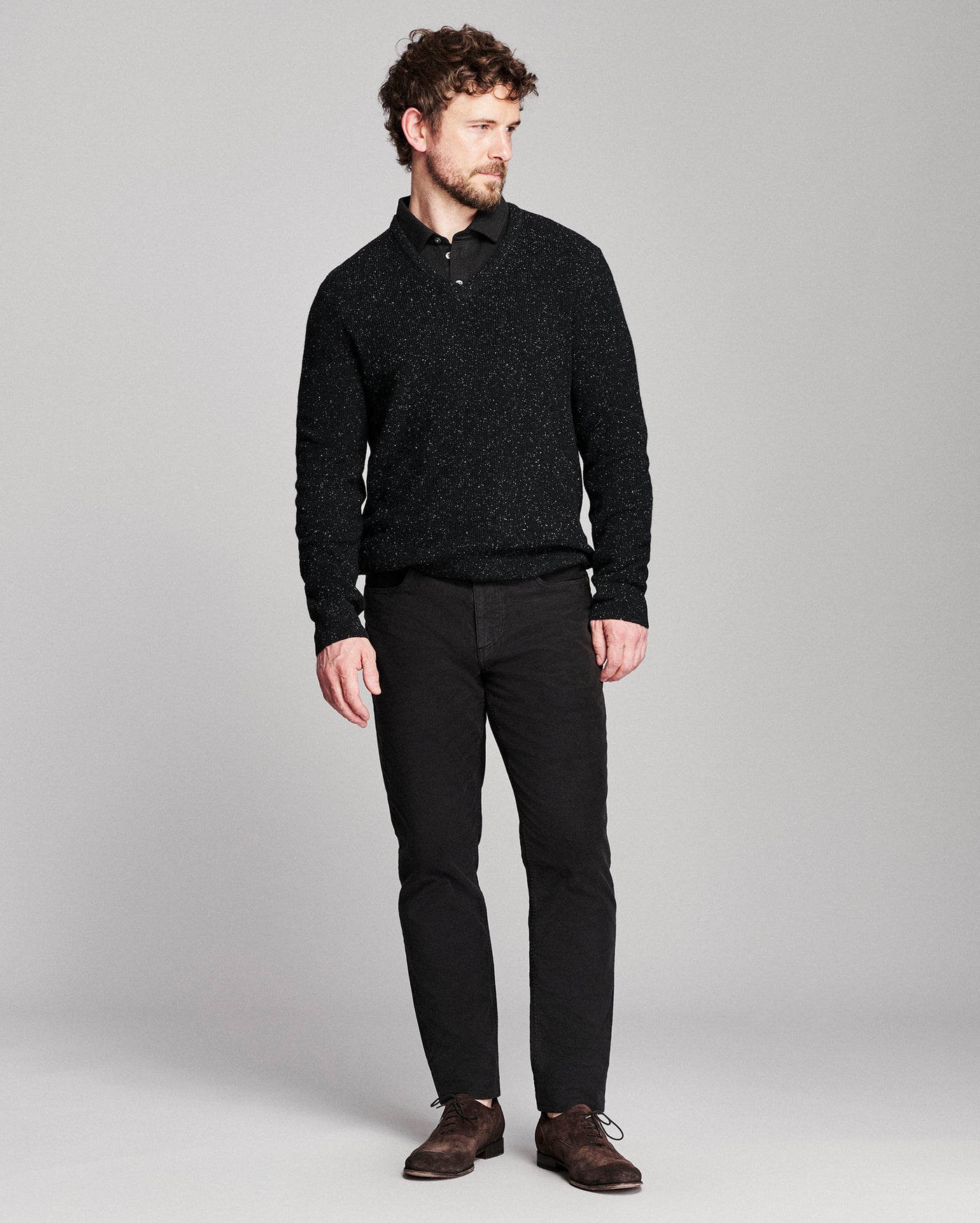 A man modeling a black textured knit polo shirt with black trousers and brown leather shoes, standing against a light gray studio background.