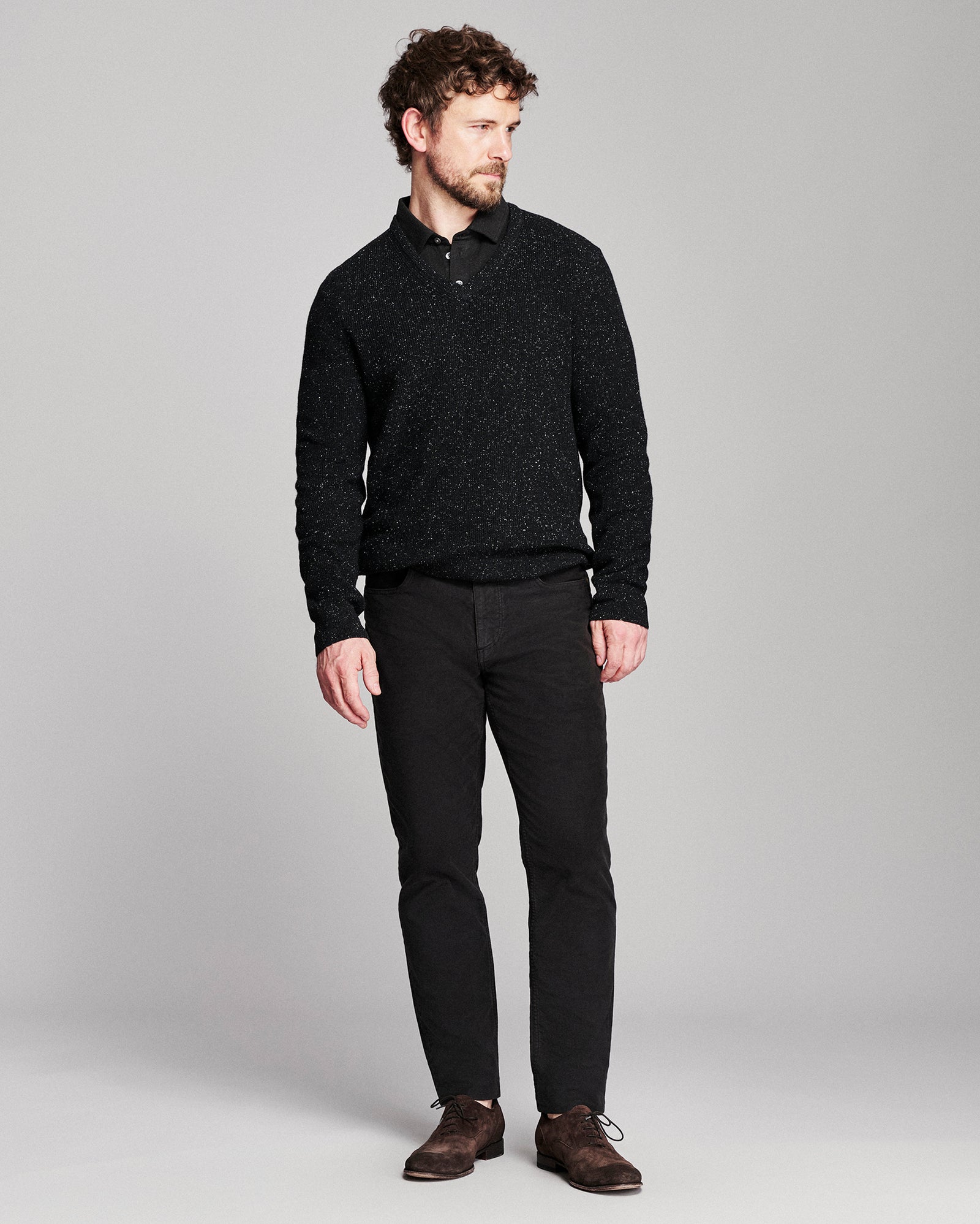 A man modeling a black textured knit polo shirt with black trousers and brown leather shoes, standing against a light gray studio background.
