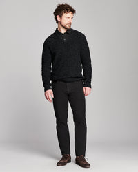 A man modeling a black textured knit polo shirt with black trousers and brown leather shoes, standing against a light gray studio background.
