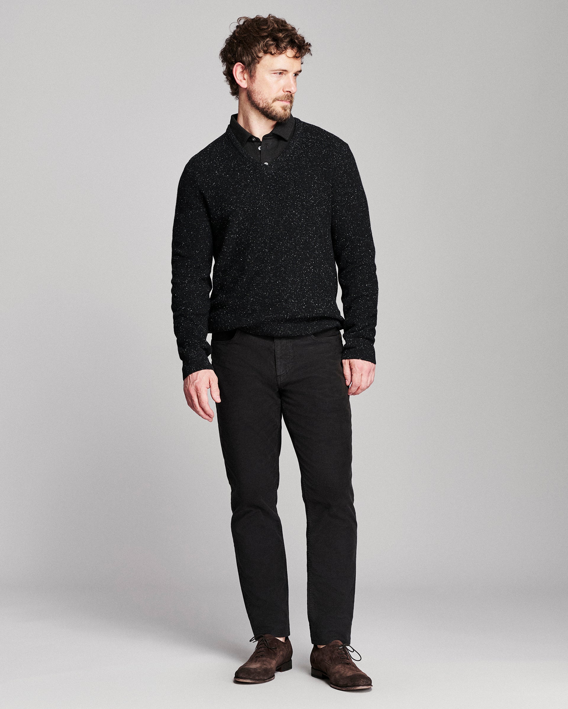 A man modeling a black textured knit polo shirt with black trousers and brown leather shoes, standing against a light gray studio background.