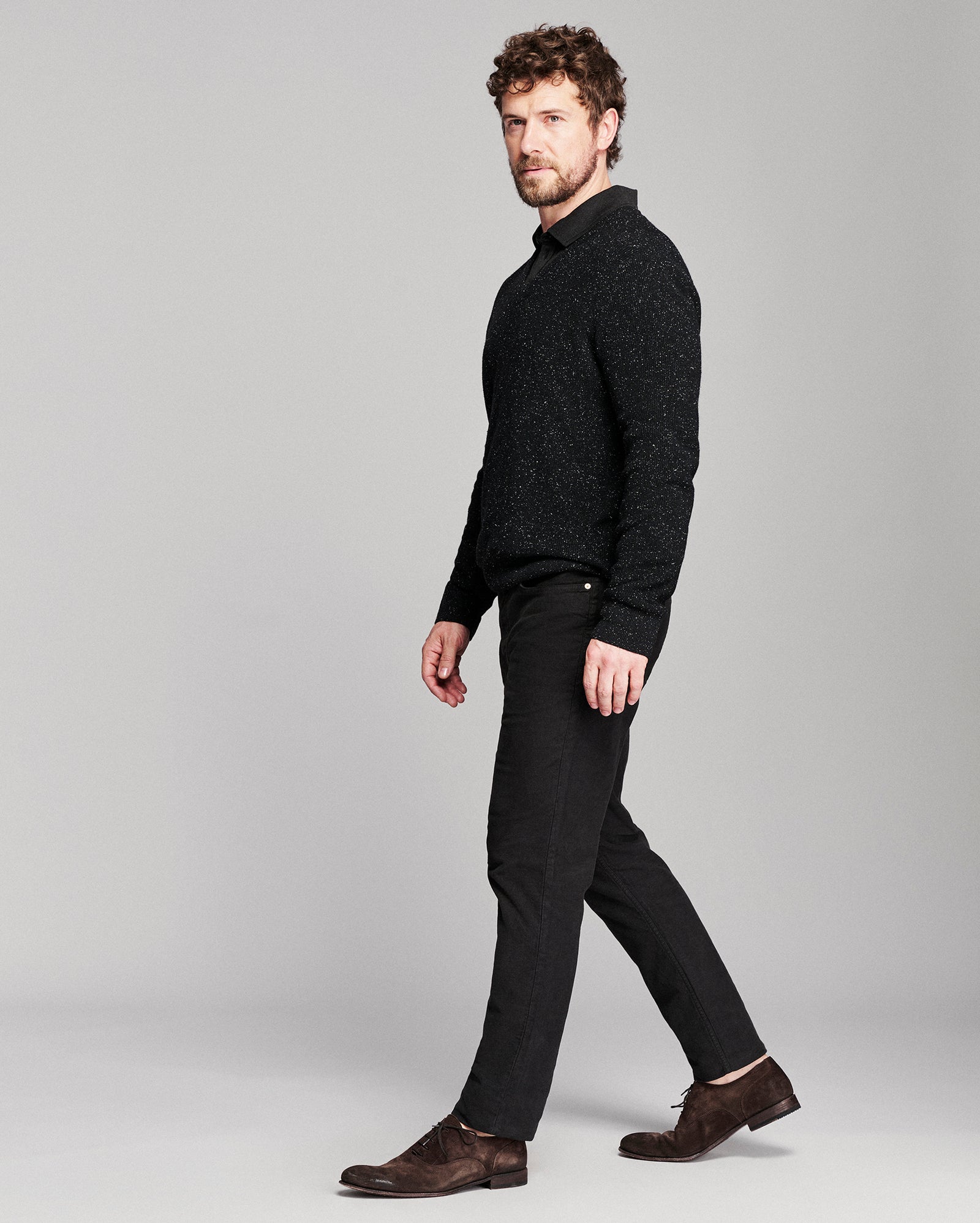 A man in a black textured knit mock-neck sweater, black trousers, and brown suede loafers walking against a light gray studio background.