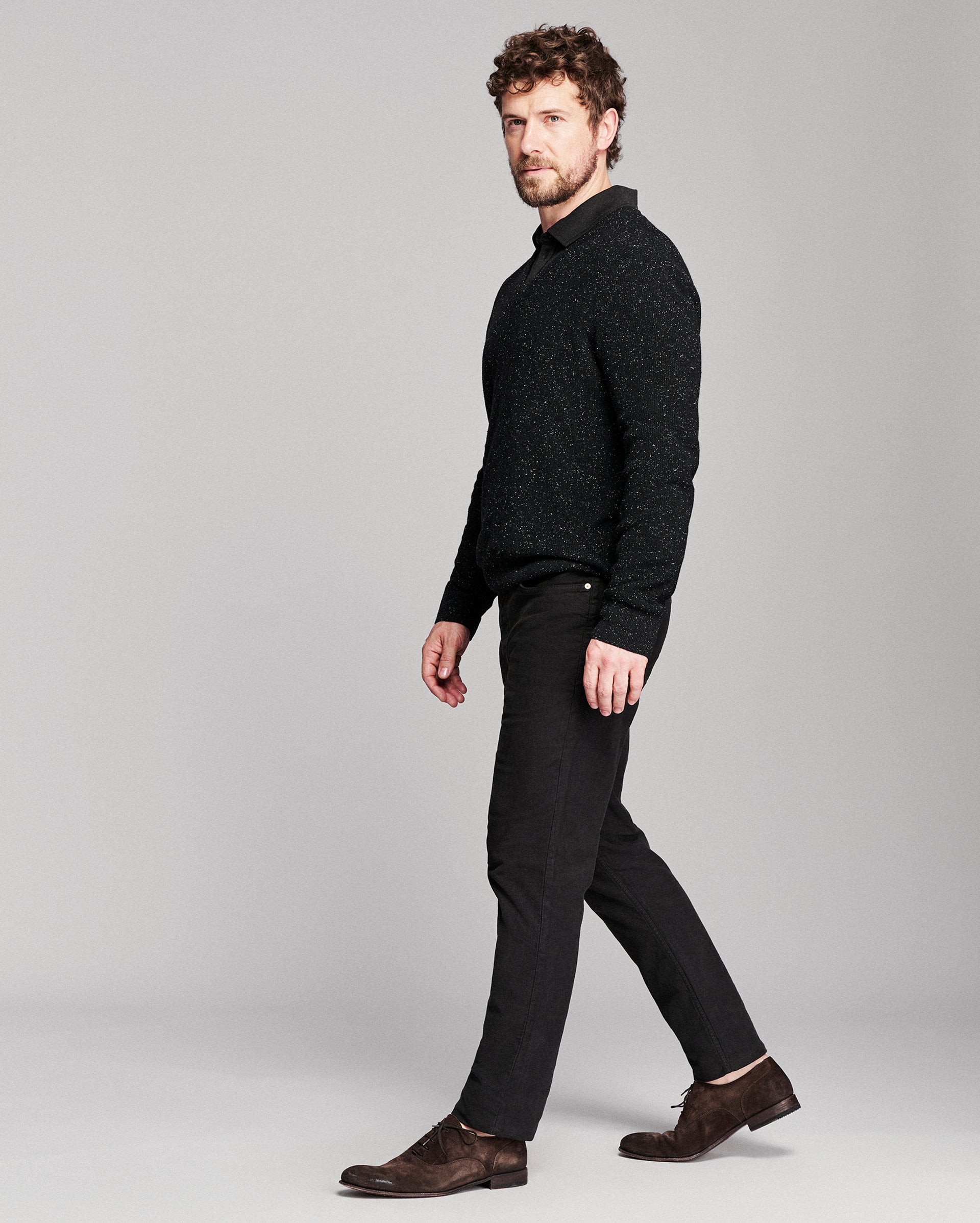 A man in a black textured knit mock-neck sweater, black trousers, and brown suede loafers walking against a light gray studio background.
