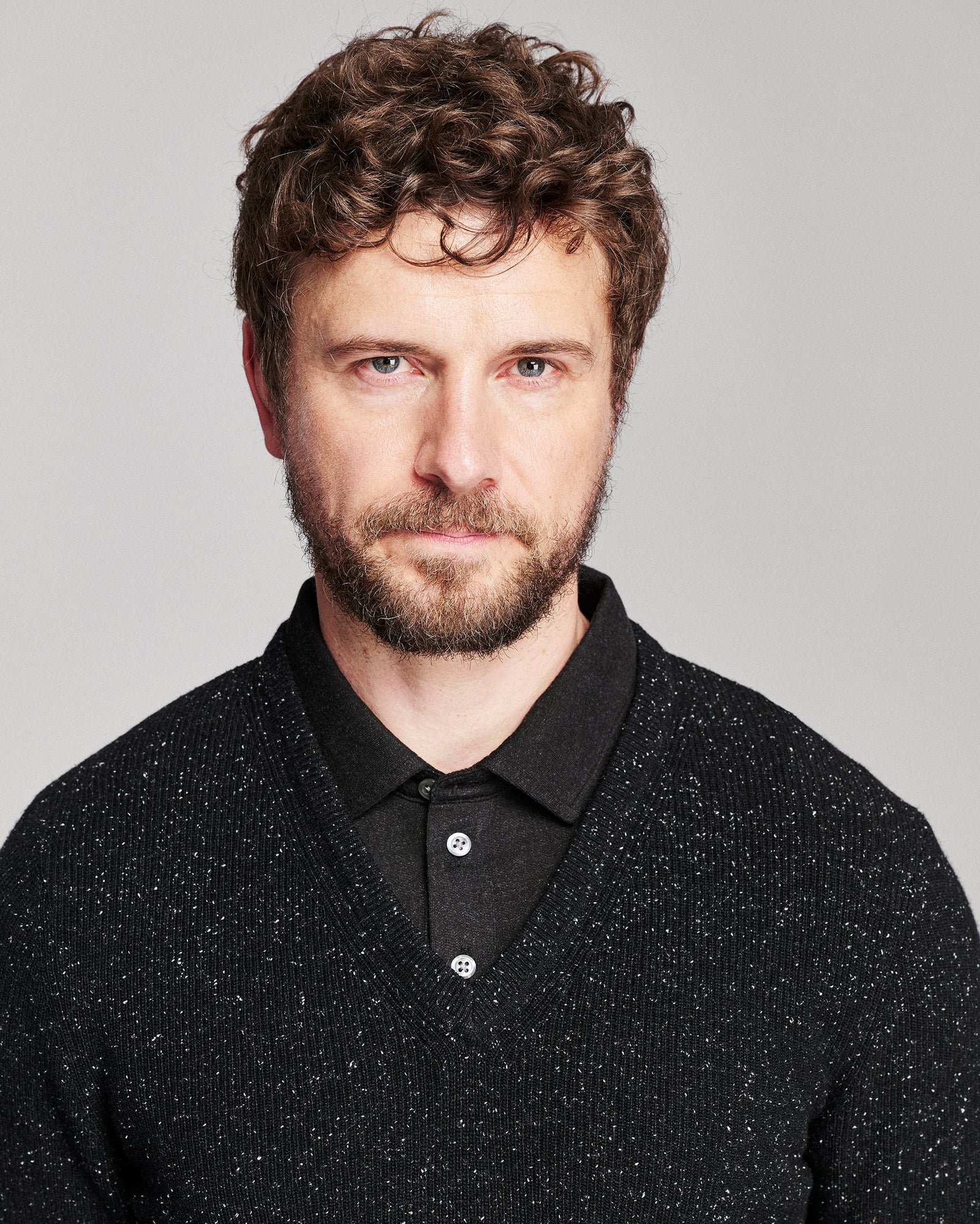 A man with curly brown hair and a beard wearing a black knit polo shirt over a black collared sweater, photographed against a light gray studio background.