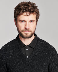 A man with curly brown hair and a beard wearing a black knit polo shirt over a black collared sweater, photographed against a light gray studio background.