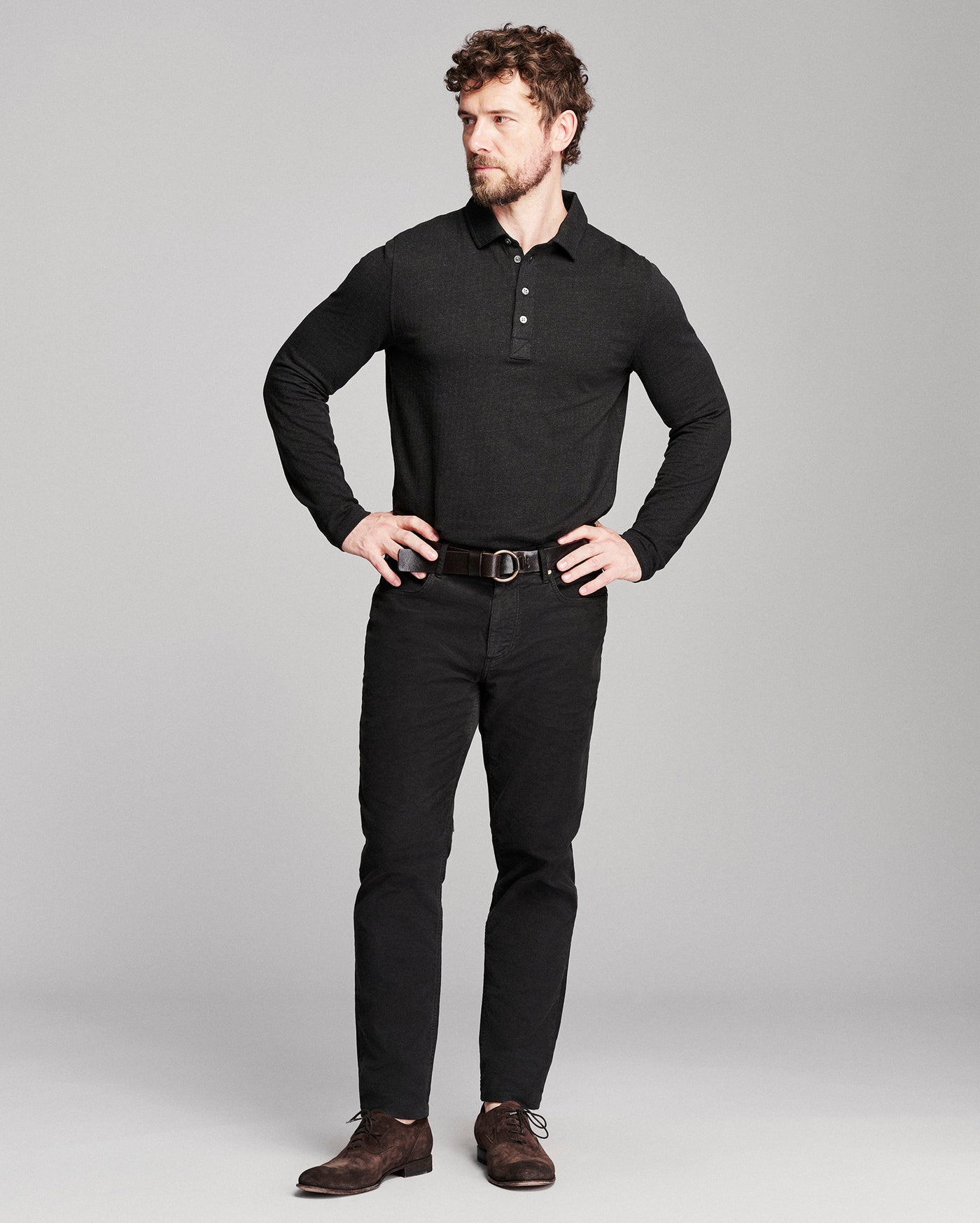 A man modeling a black long-sleeve polo shirt, black pants, a dark brown belt, and brown suede shoes against a light gray studio background.