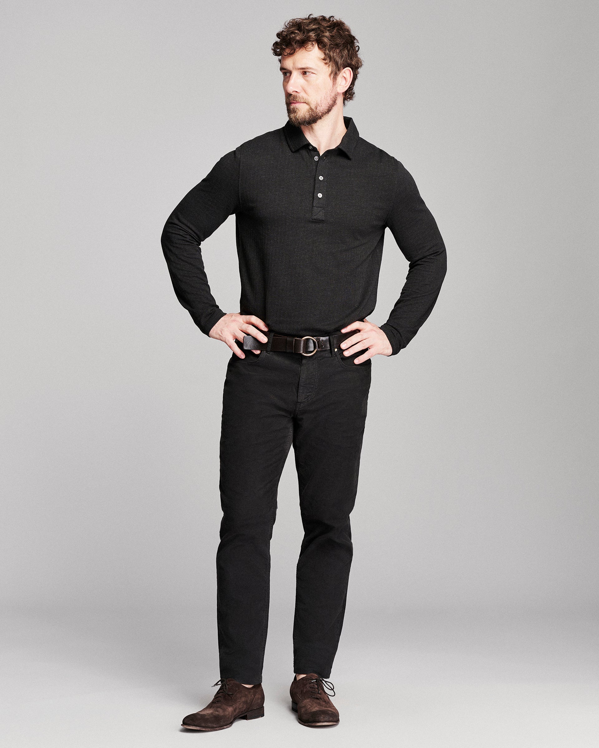 A man modeling a black long-sleeve polo shirt, black pants, a dark brown belt, and brown suede shoes against a light gray studio background.