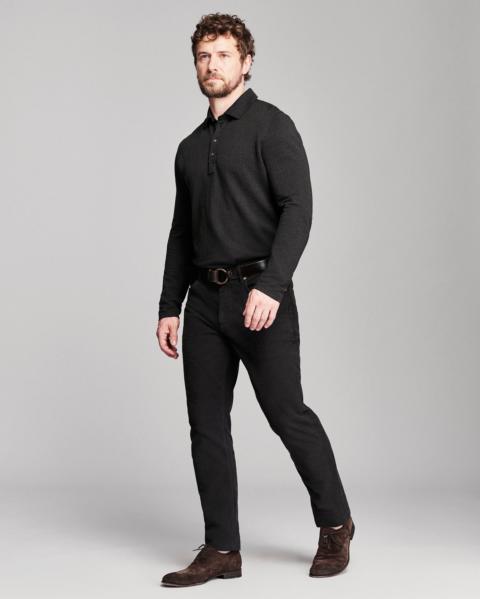 A man modeling a black long-sleeve knit polo shirt, black trousers, a black belt, and brown suede shoes against a solid gray studio background.