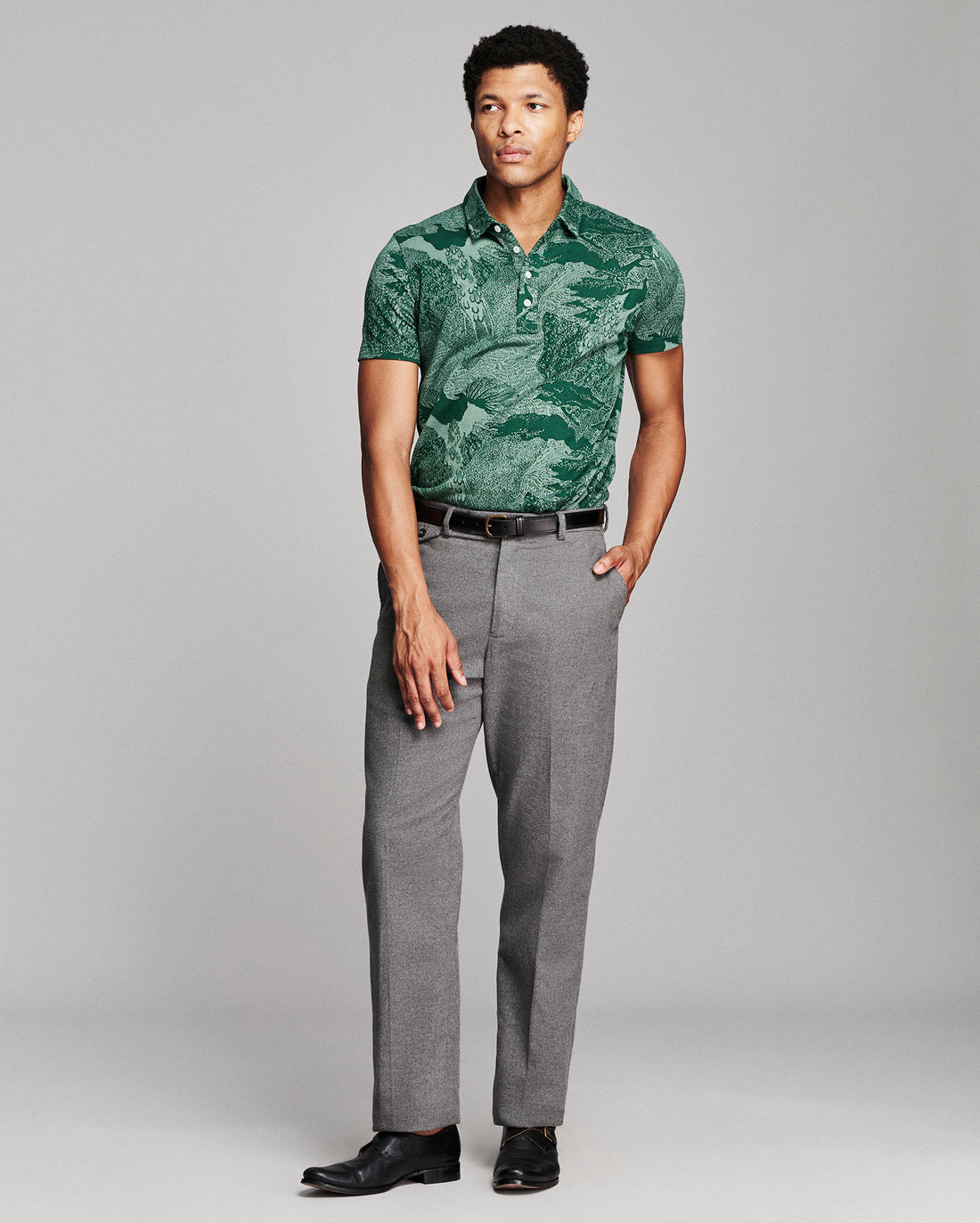 A man modeling a green tropical leaf print short-sleeve polo shirt paired with gray trousers and black dress shoes, standing against a light gray studio background.