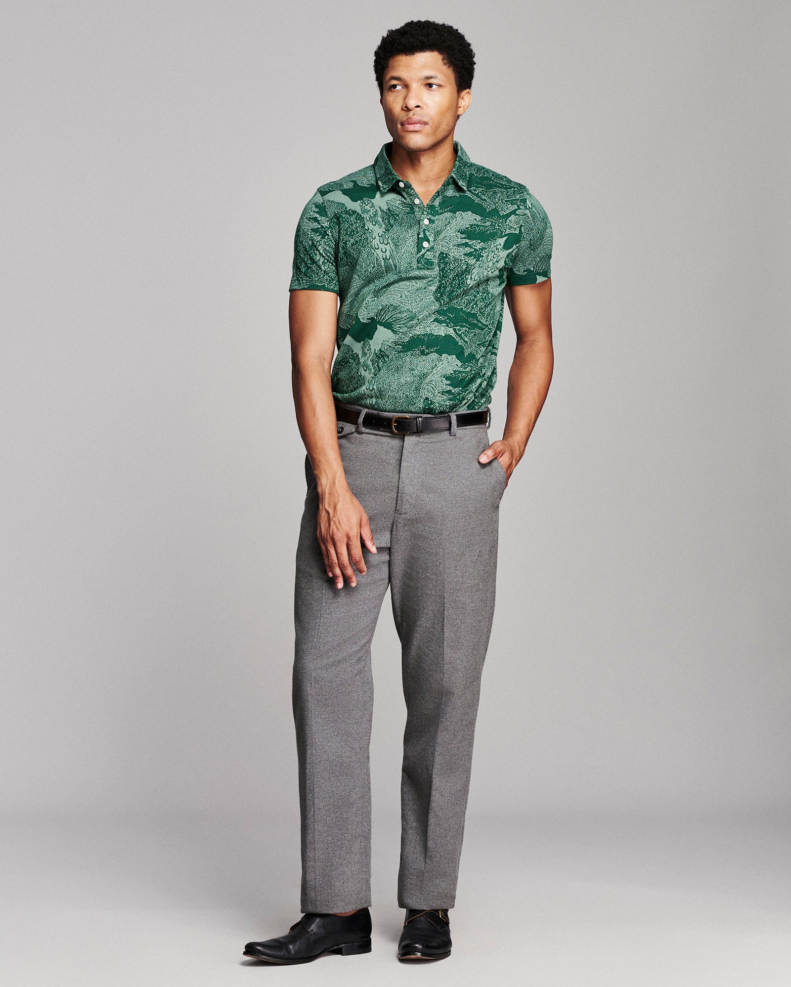 A man modeling a green tropical leaf print short-sleeve polo shirt paired with gray trousers and black dress shoes, standing against a light gray studio background.