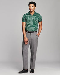 A man modeling a green tropical leaf print short-sleeve polo shirt paired with gray trousers and black dress shoes, standing against a light gray studio background.