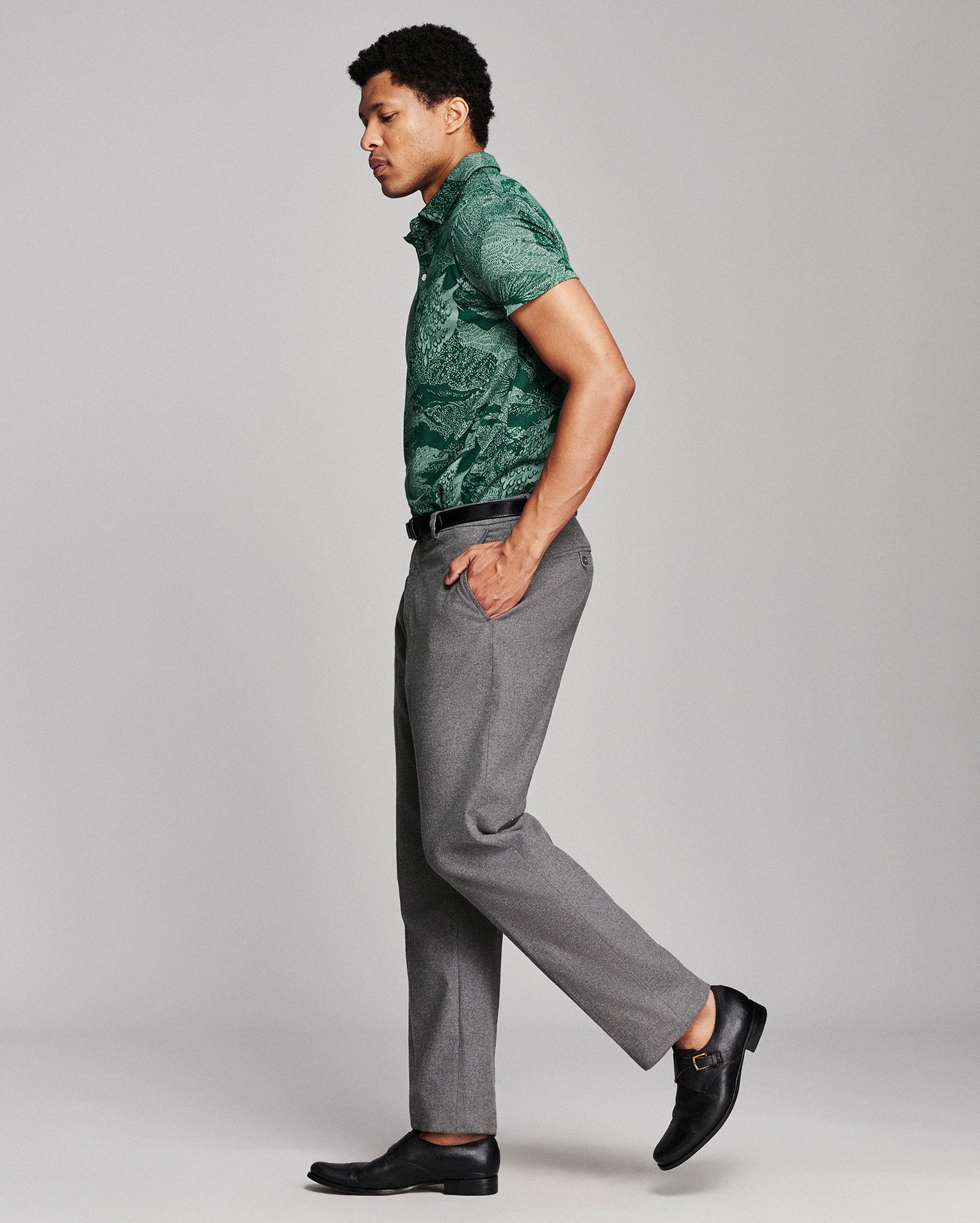 A man in side profile wearing a green short-sleeve patterned polo shirt, gray trousers, and black loafers, standing against a light gray studio background.