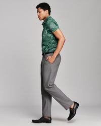 A man in side profile wearing a green short-sleeve patterned polo shirt, gray trousers, and black loafers, standing against a light gray studio background.