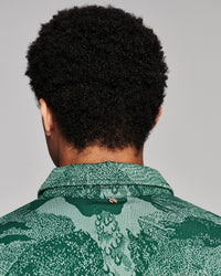 Back view of a person with curly black hair wearing a green patterned collared shirt against a light gray studio background.