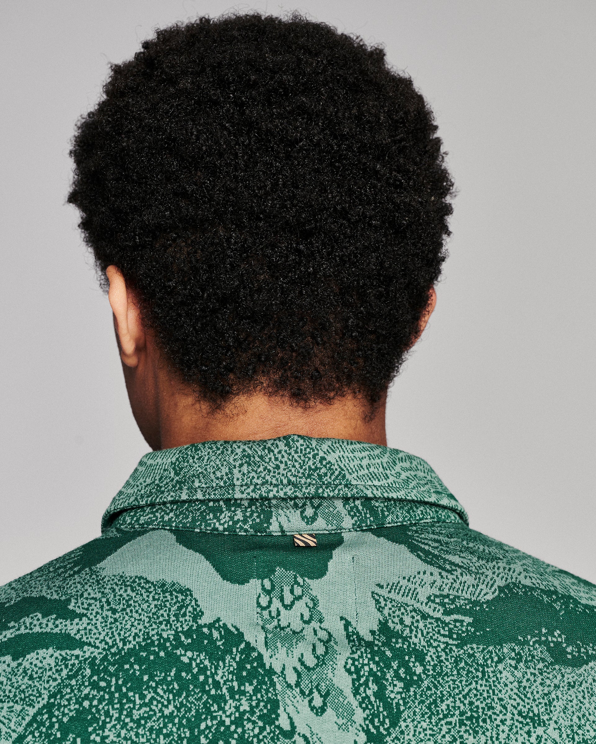 Back view of a person with curly black hair wearing a green patterned collared shirt against a light gray studio background.