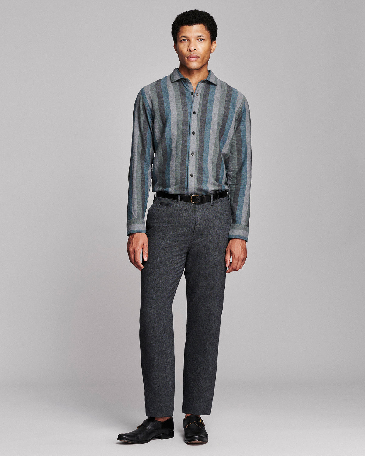 A man modeling a long-sleeve button-up shirt with vertical blue and gray stripes, paired with dark gray trousers, a black belt, and black dress shoes, against a light gray studio background.