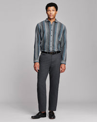 A man modeling a long-sleeve button-up shirt with vertical blue and gray stripes, paired with dark gray trousers, a black belt, and black dress shoes, against a light gray studio background.