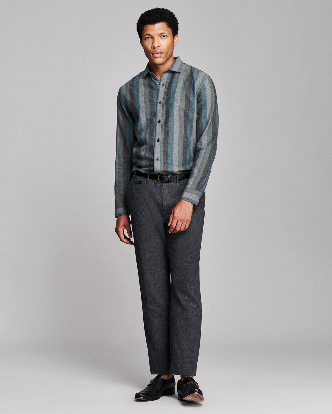 A man modeling a long-sleeve button-up shirt with vertical blue and gray stripes, paired with dark gray trousers and black shoes, against a light gray studio background.