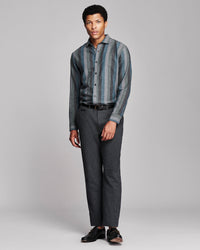 A man modeling a long-sleeve button-up shirt with vertical blue and gray stripes, paired with dark gray trousers and black shoes, against a light gray studio background.